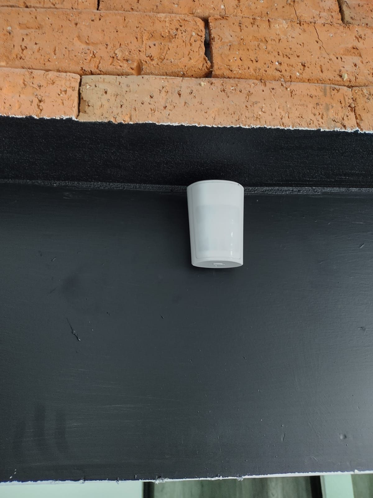 Cámara de seguridad blanca montada en una repisa pintada de negro debajo de una pared de ladrillos.