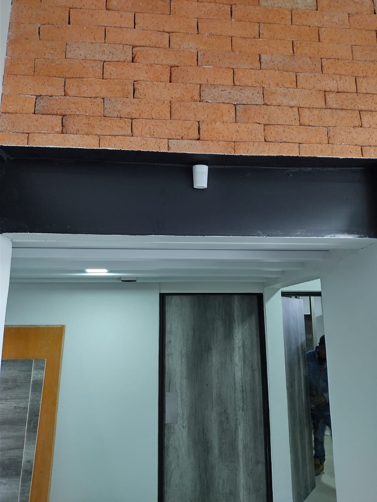 Una puerta de entrada a una habitación con una viga negra, una pared de ladrillos encima y una lámpara cilíndrica blanca.