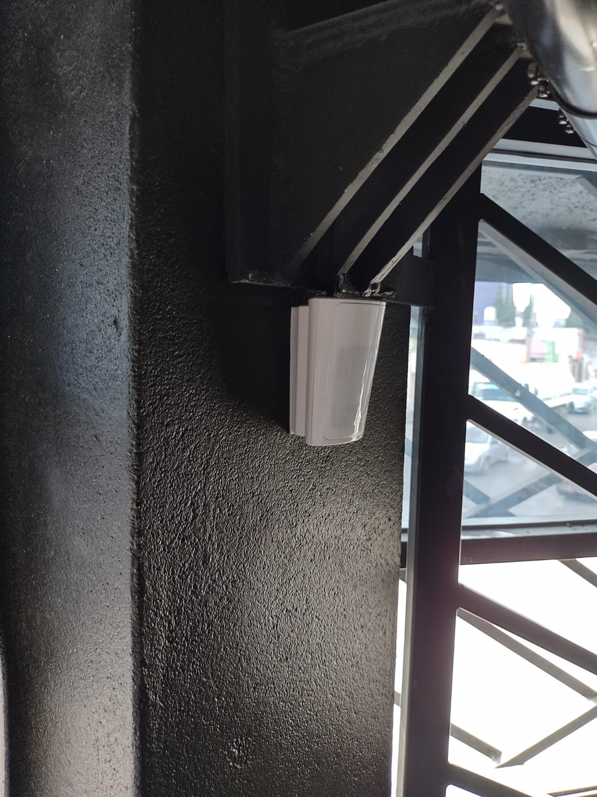 Altavoz blanco montado en una pared negra texturizada, cerca de una ventana con una viga de soporte de metal.