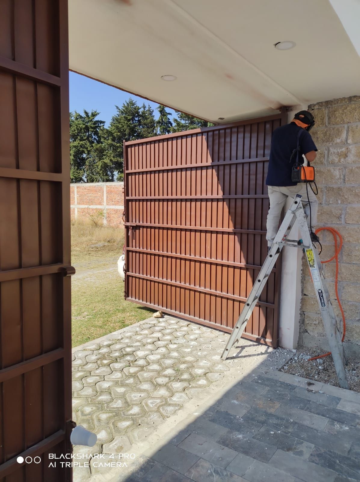 Persona en una escalera, cortando una puerta metálica marrón cerca de la entrada de una casa. Luz solar, exterior.