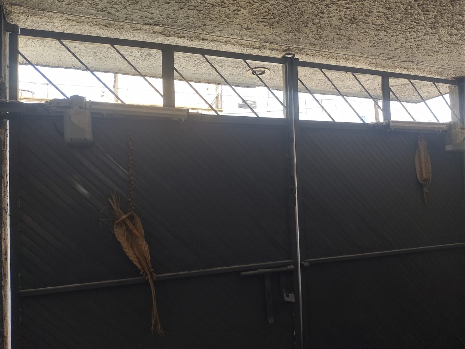 Una puerta de metal con una ventana con diseño de rombos en la parte superior. Dos plantas secas cuelgan de la puerta.