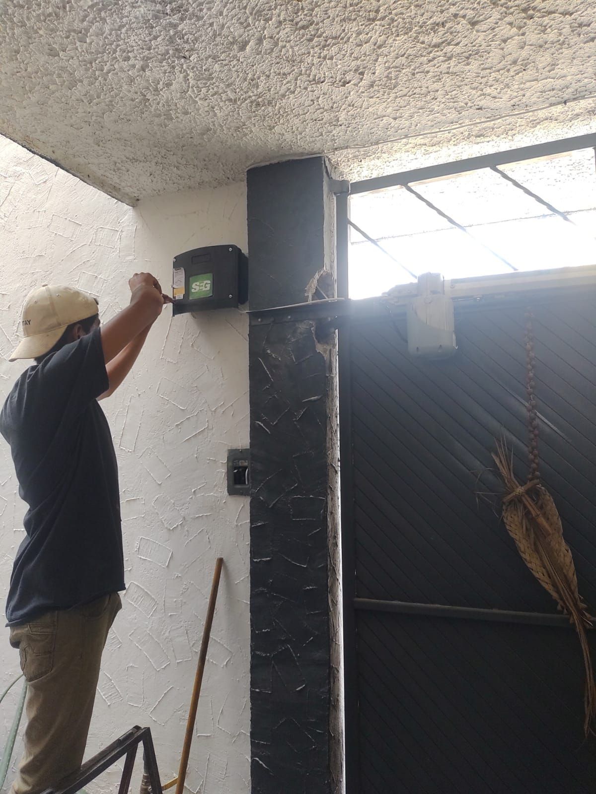 Hombre en una escalera instalando un abridor de puerta en un pilar negro al lado de una puerta de metal negra.