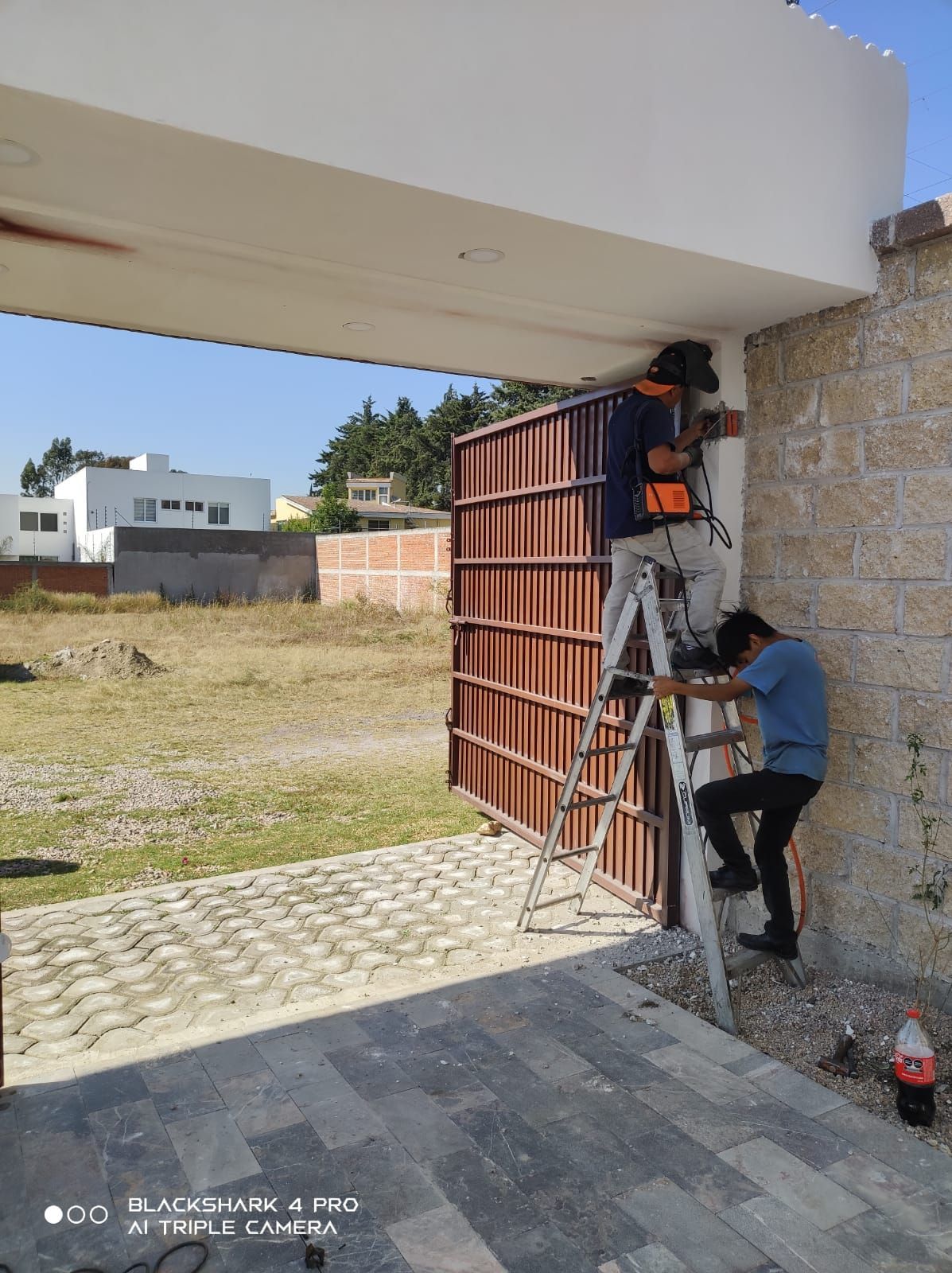 Dos hombres instalando una cámara de seguridad en un pilar blanco junto a una puerta de metal marrón, al aire libre.