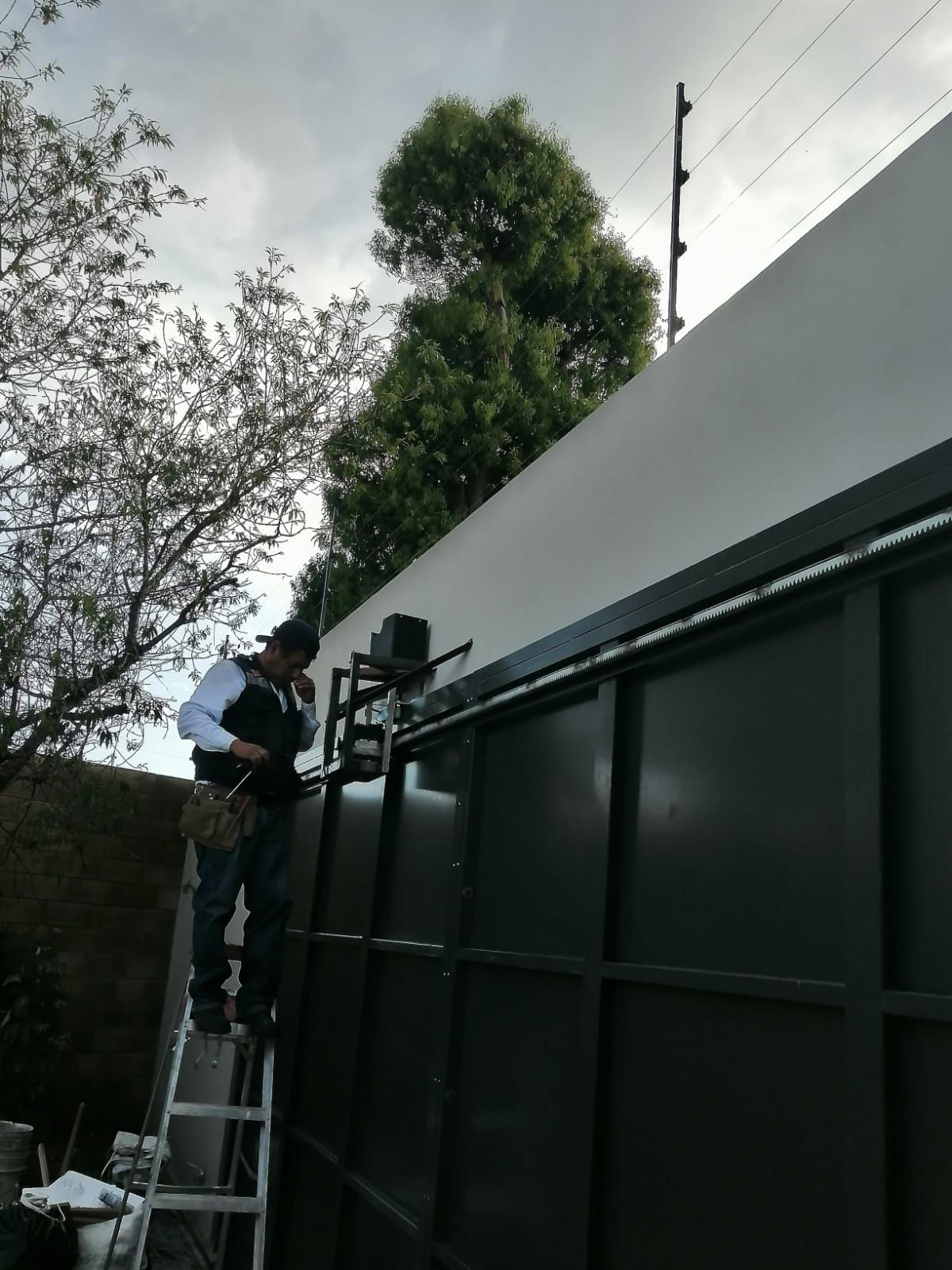 Un hombre en una escalera instala un sistema de seguridad en un edificio verde oscuro; hay una cerca de alambre de púas en el techo.