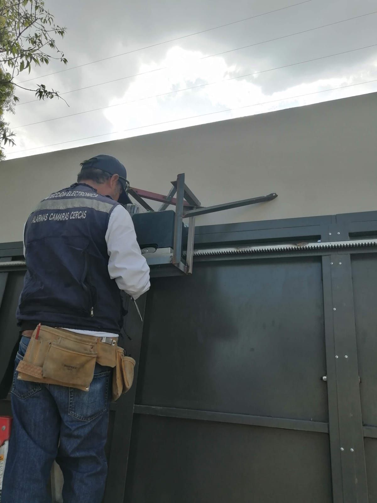 Un hombre con chaleco trabaja en una puerta automática con herramientas. Pared beige y cielo nublado al fondo.