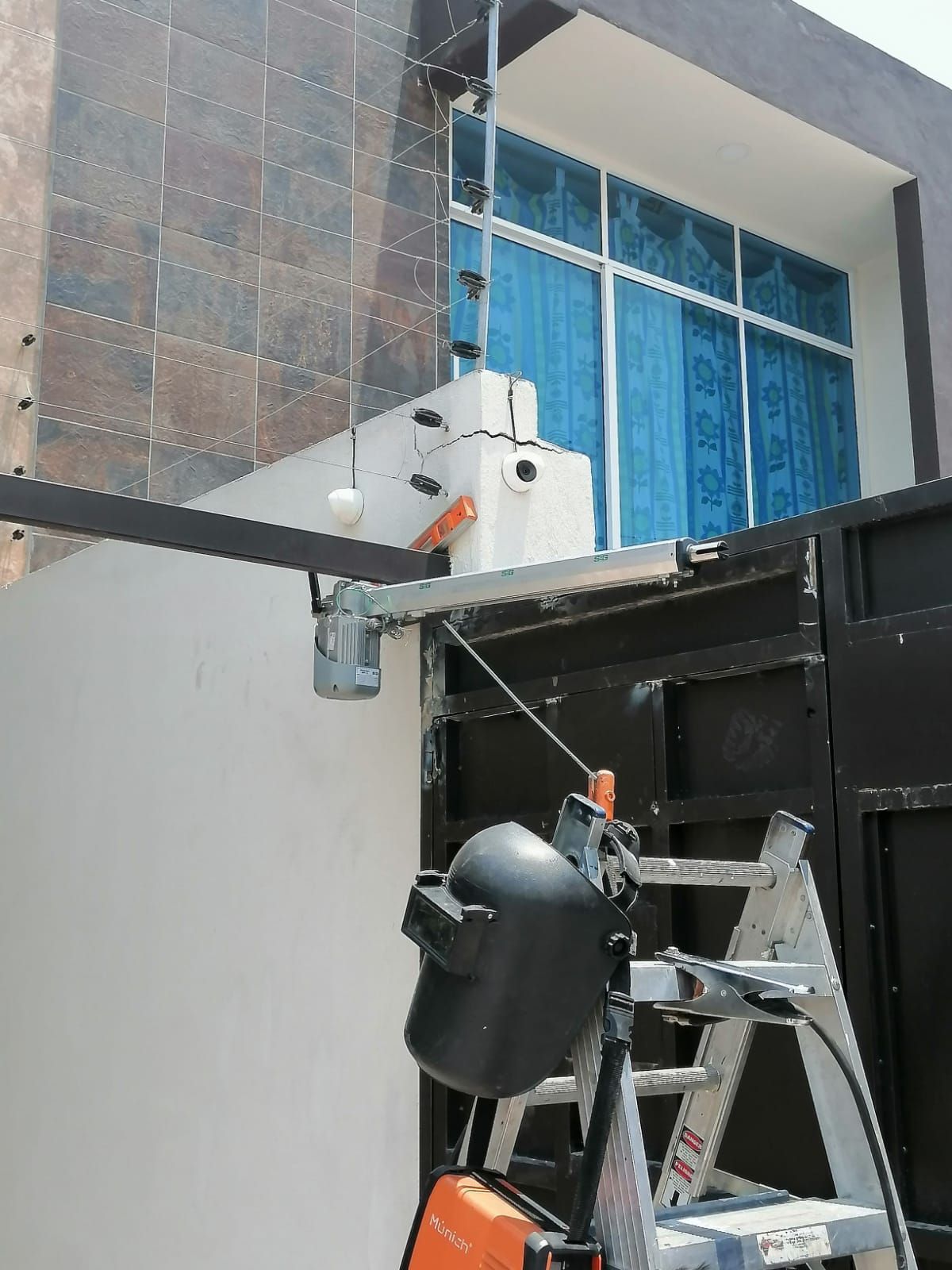 Soldador trabajando en un portón eléctrico, con escalera, fachada de edificio y sistema de seguridad.