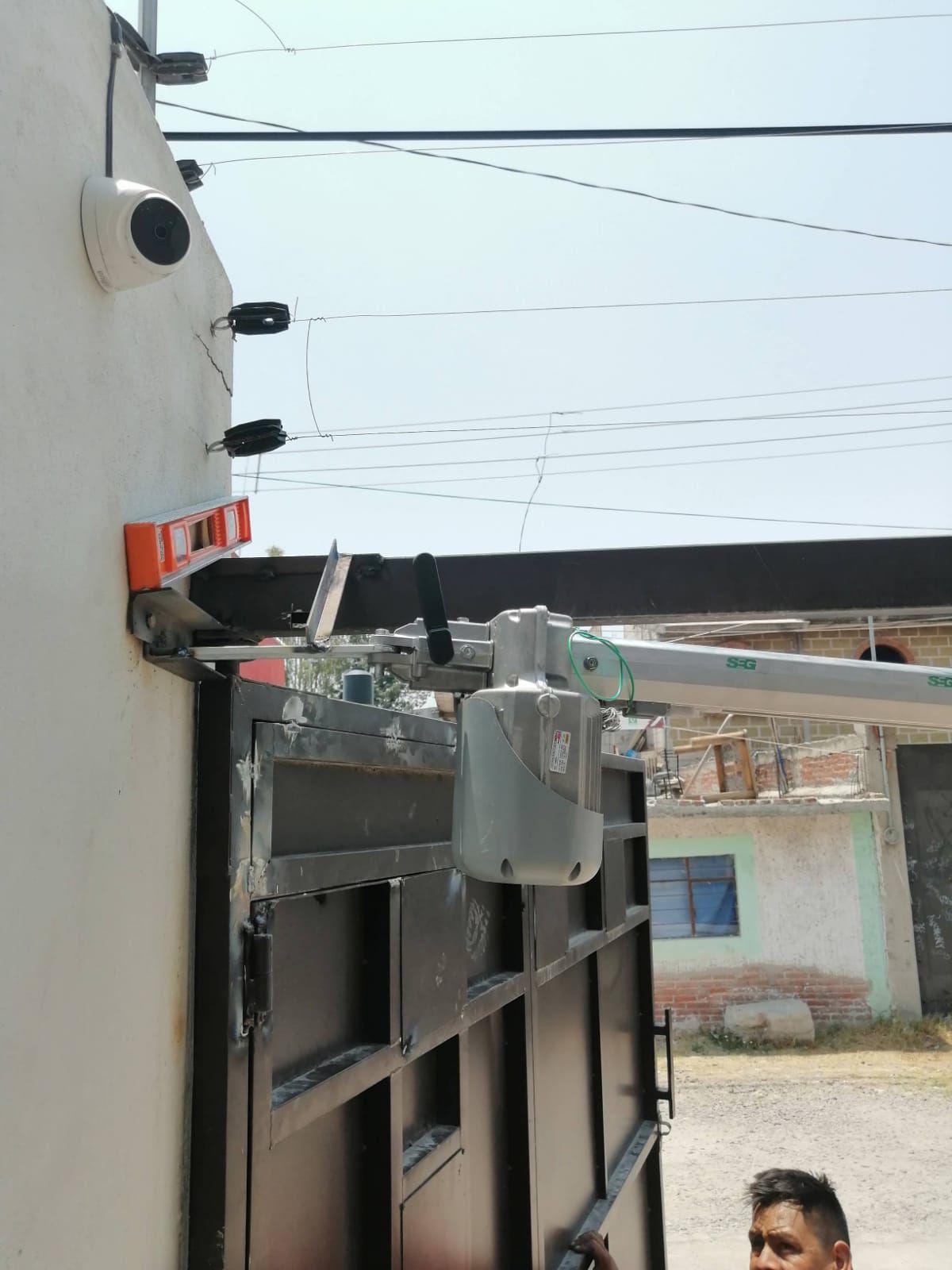 Portón automático con motor, cámara de seguridad y cerca eléctrica en una pared blanca. Luminoso espacio al aire libre.