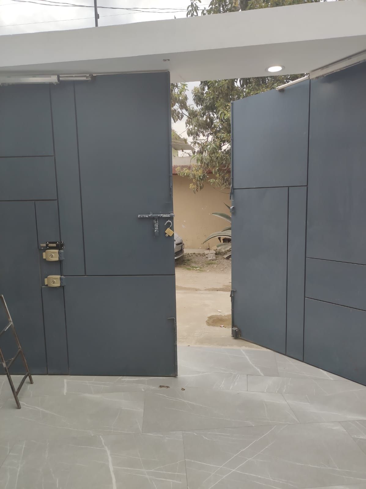 Puerta metálica gris, parcialmente abierta, que deja ver un patio. Luz solar.