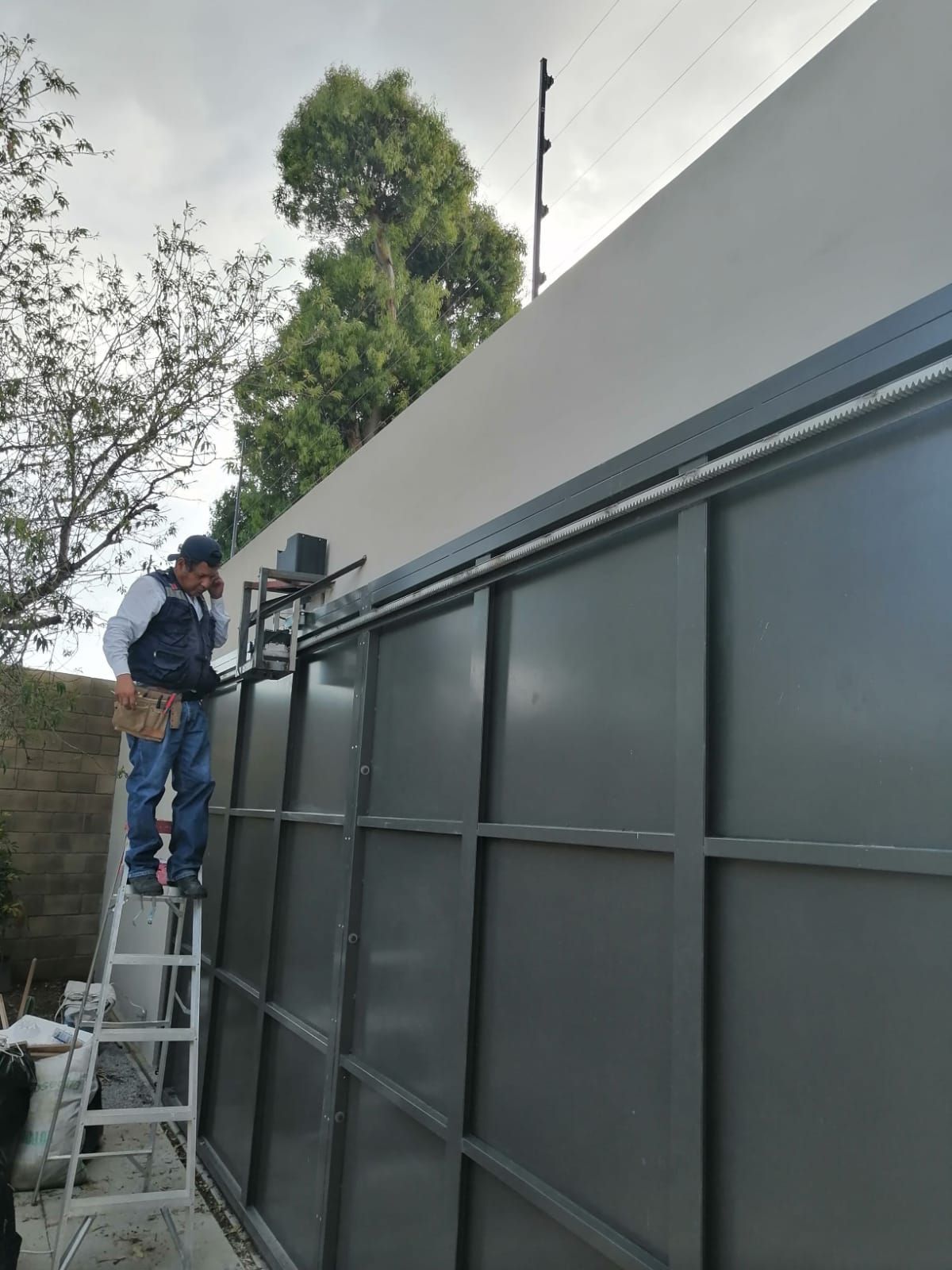 Hombre en una escalera instalando una puerta corrediza con una cerca eléctrica encima. Paredes grises y blancas.