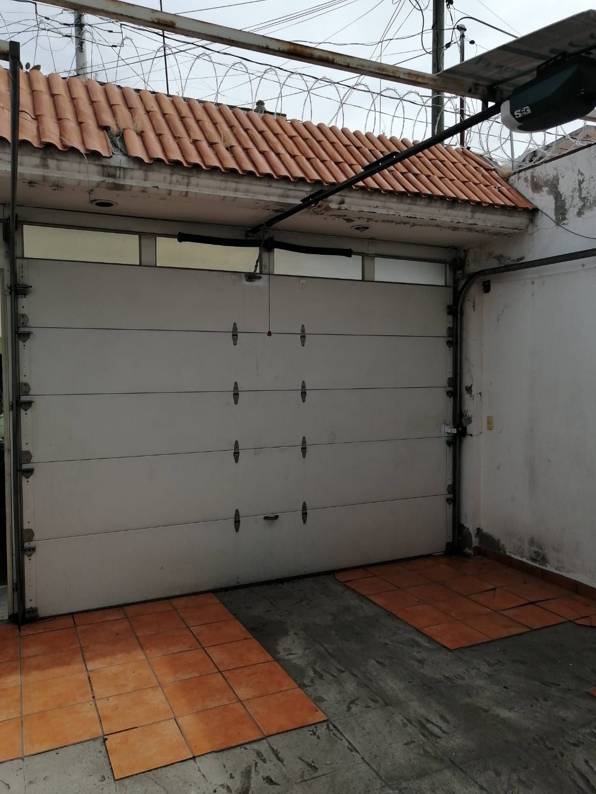 Puerta de garaje blanca cerrada bajo un techo de tejas naranjas. Cerca de alambre de púas encima.