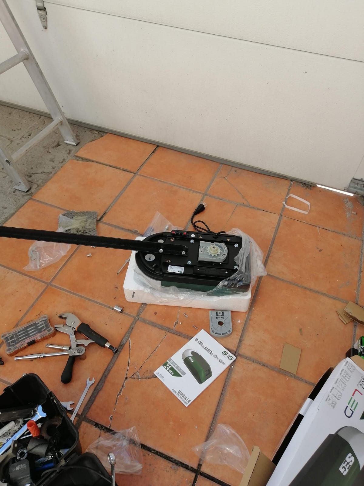 Piezas del abridor de puertas de garaje dispuestas sobre un piso de baldosas junto a una pared blanca.