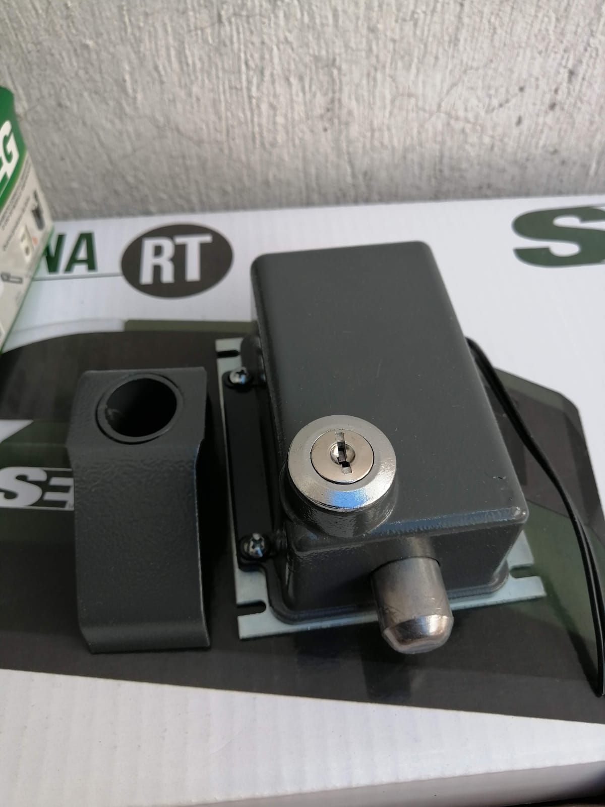 Cerradura de metal gris con cerradura redonda y extremo receptor negro a juego, sobre una superficie rayada.