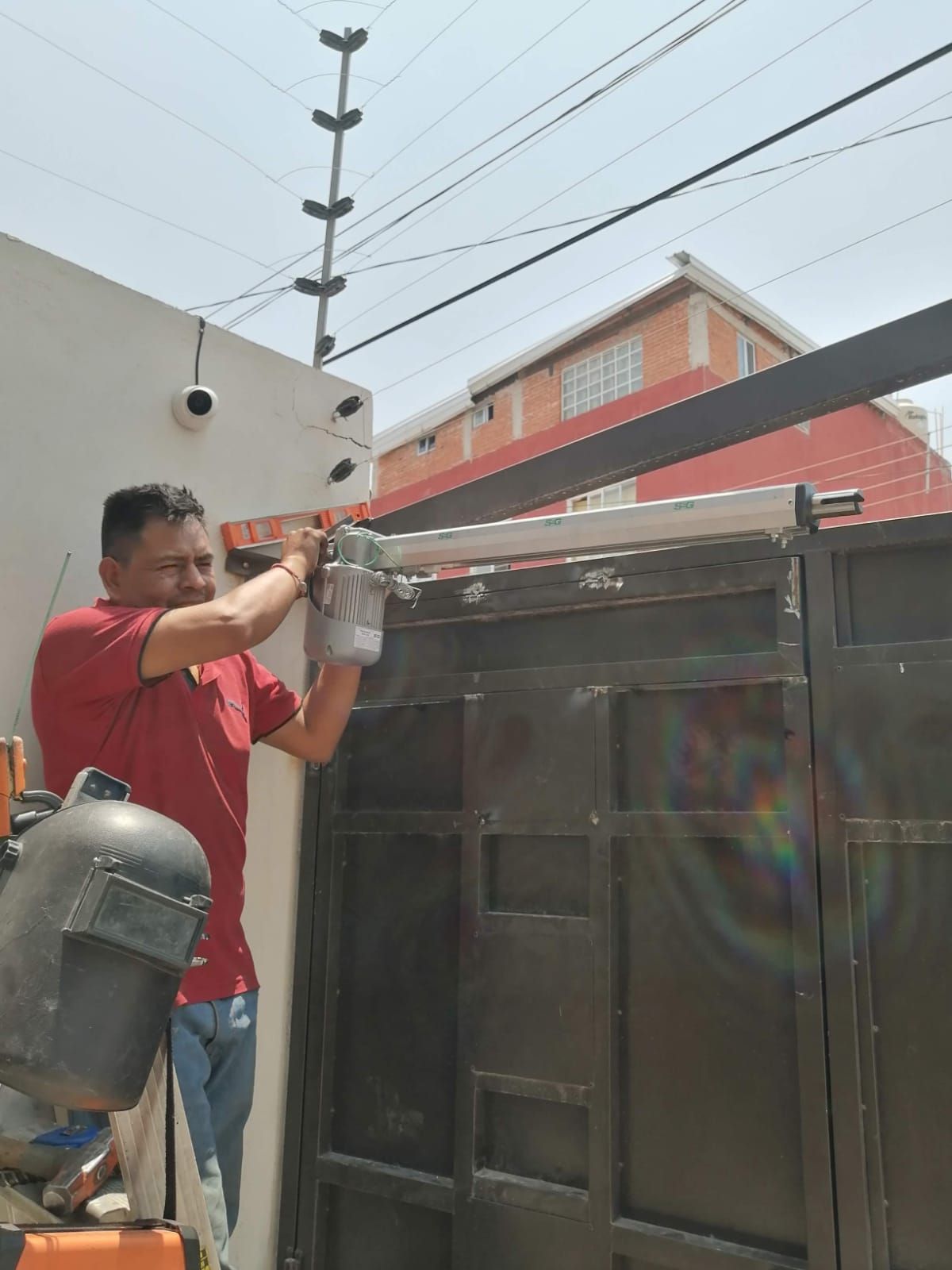 Hombre instalando un abridor de puerta en una puerta de metal; valla de seguridad y edificio al fondo.