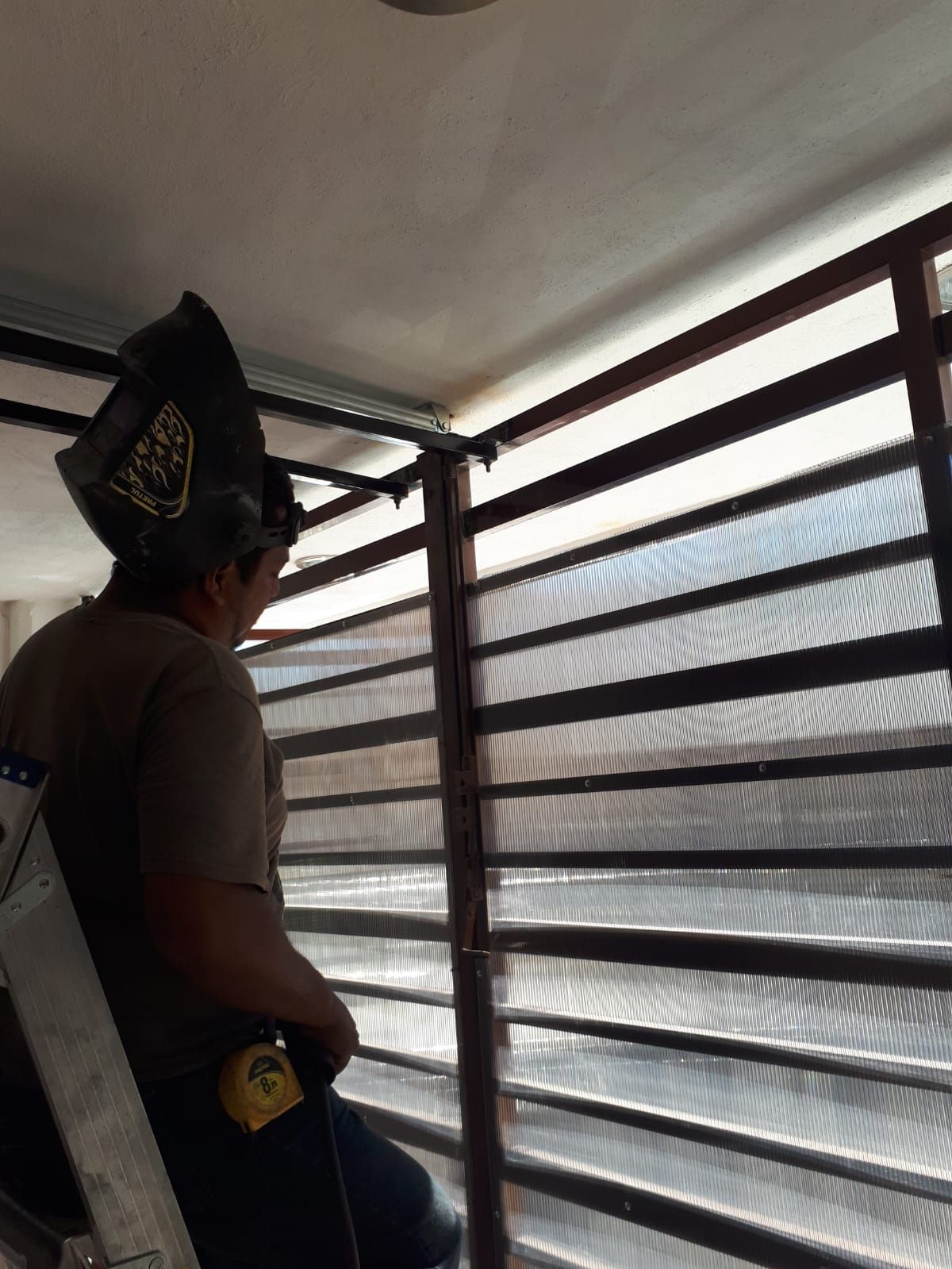Una persona con casco instala persianas, en el interior, al lado de una ventana.