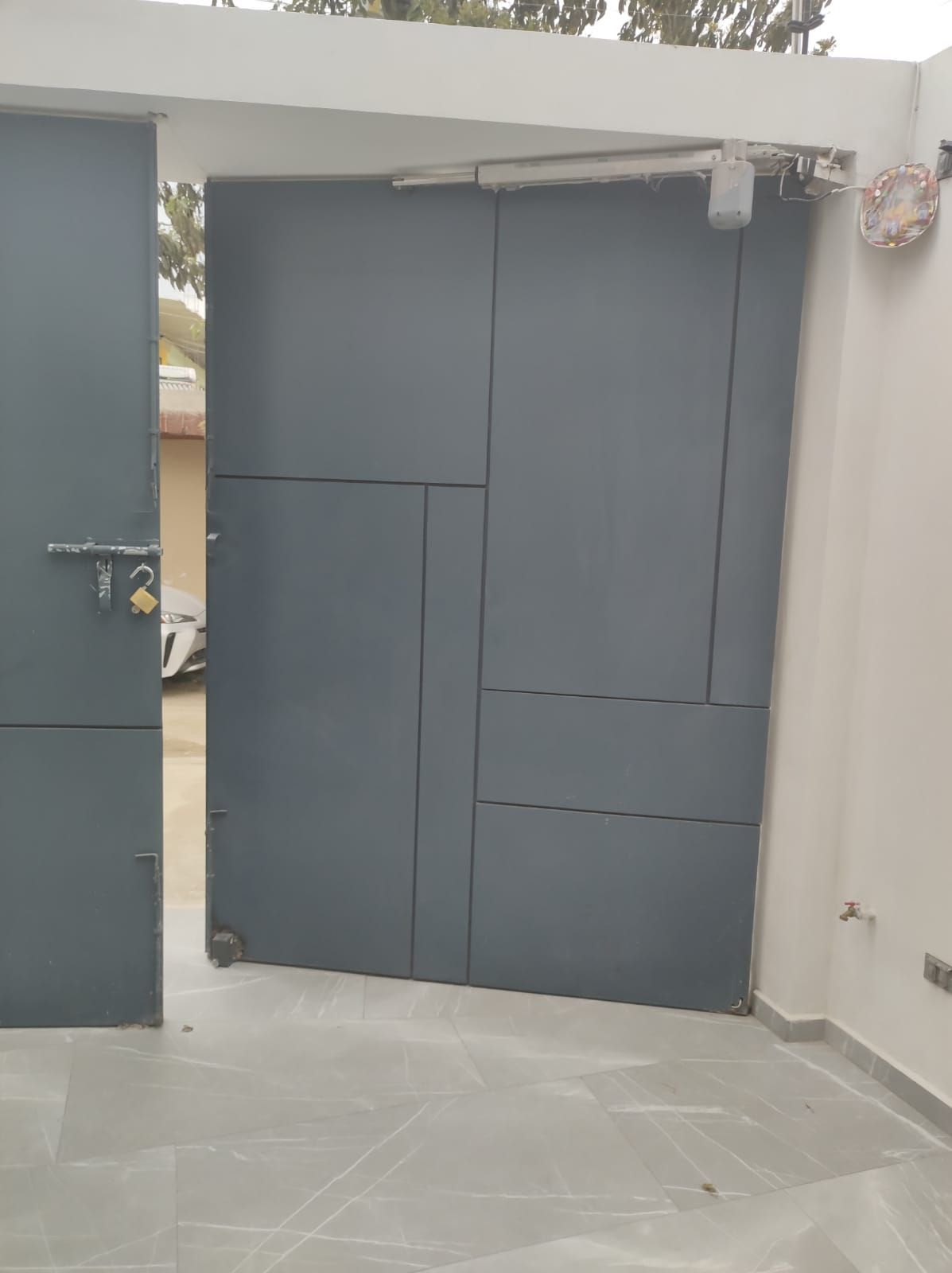 Una puerta de metal gris, parcialmente abierta, con abridor automático, ubicada en un área pavimentada.