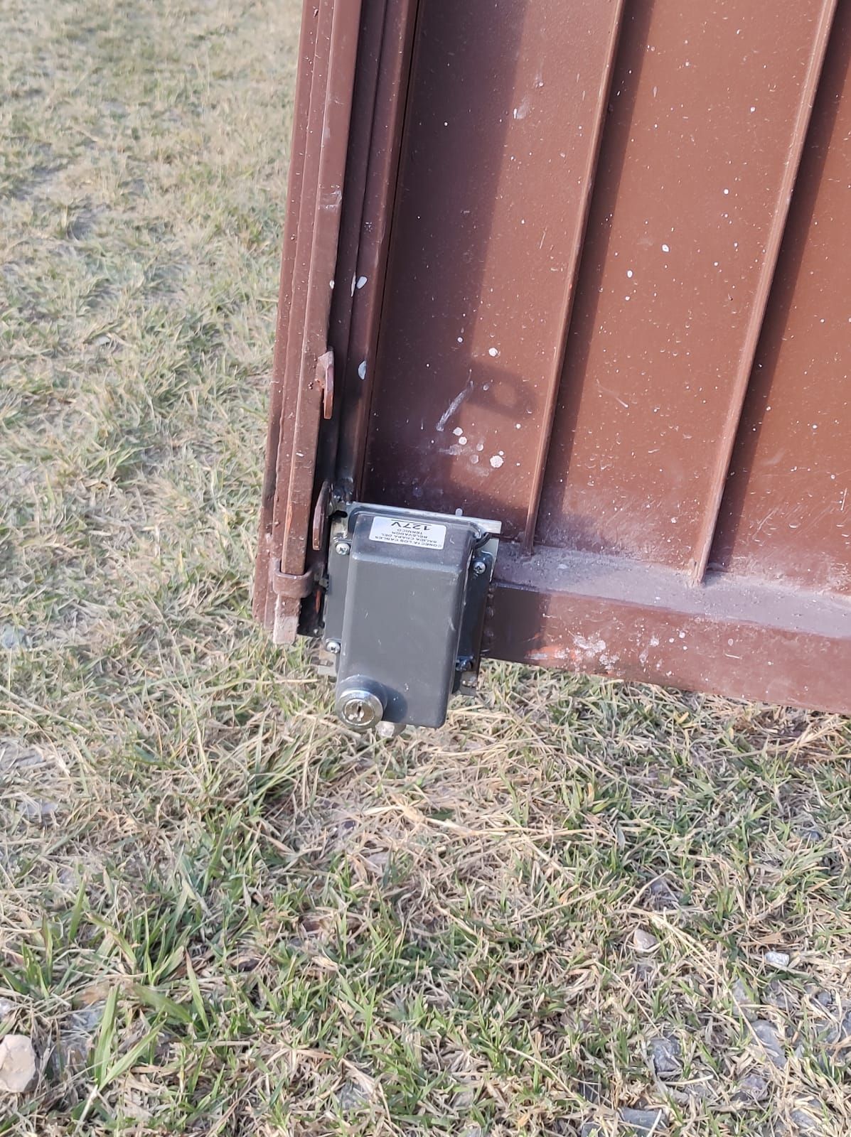 Puerta metálica de color marrón con dispositivo rectangular fijado a nivel del suelo, situada sobre césped.