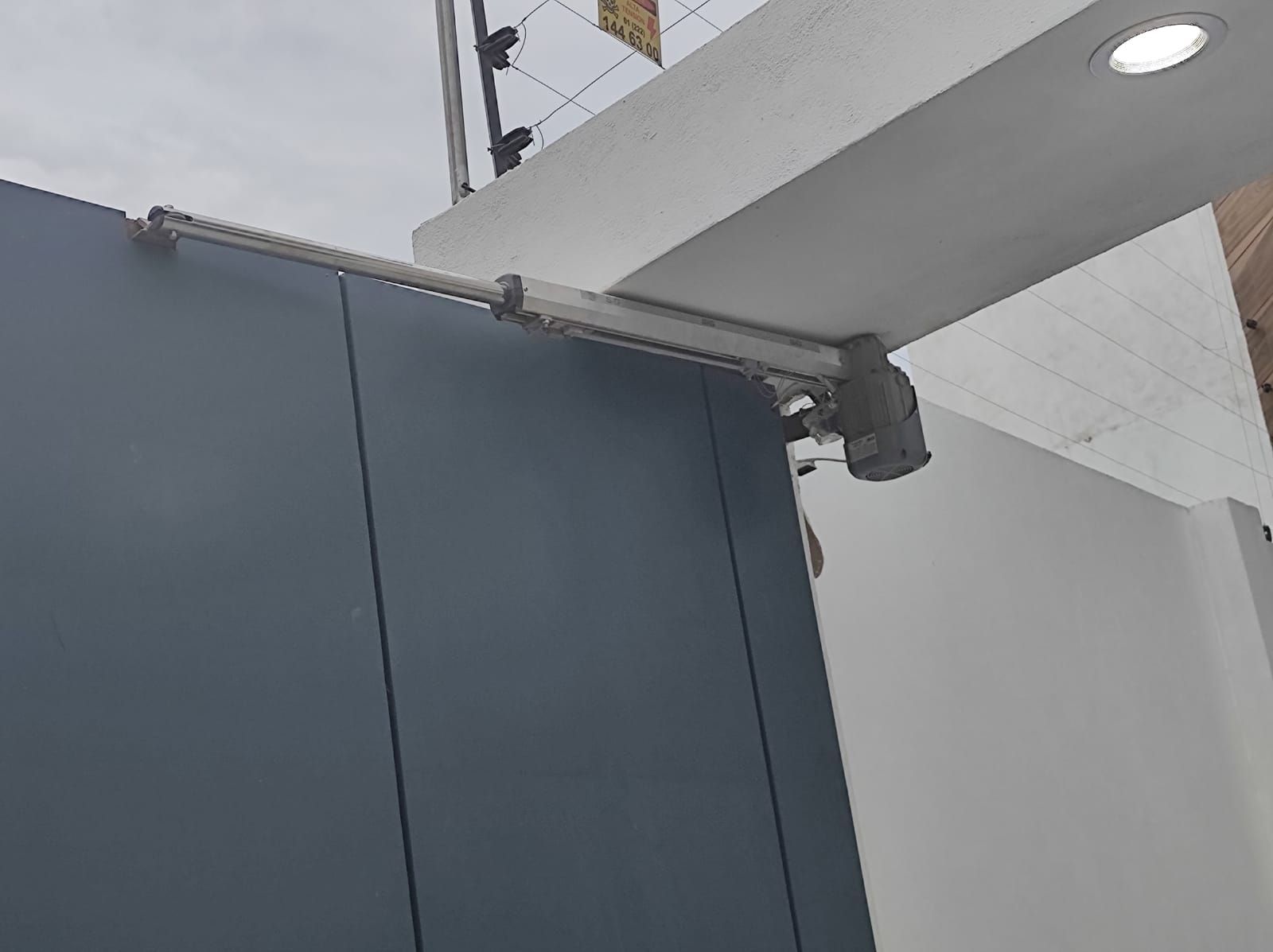 Una puerta azul con un abridor de brazo automático unido a una pared blanca, en un entorno al aire libre.