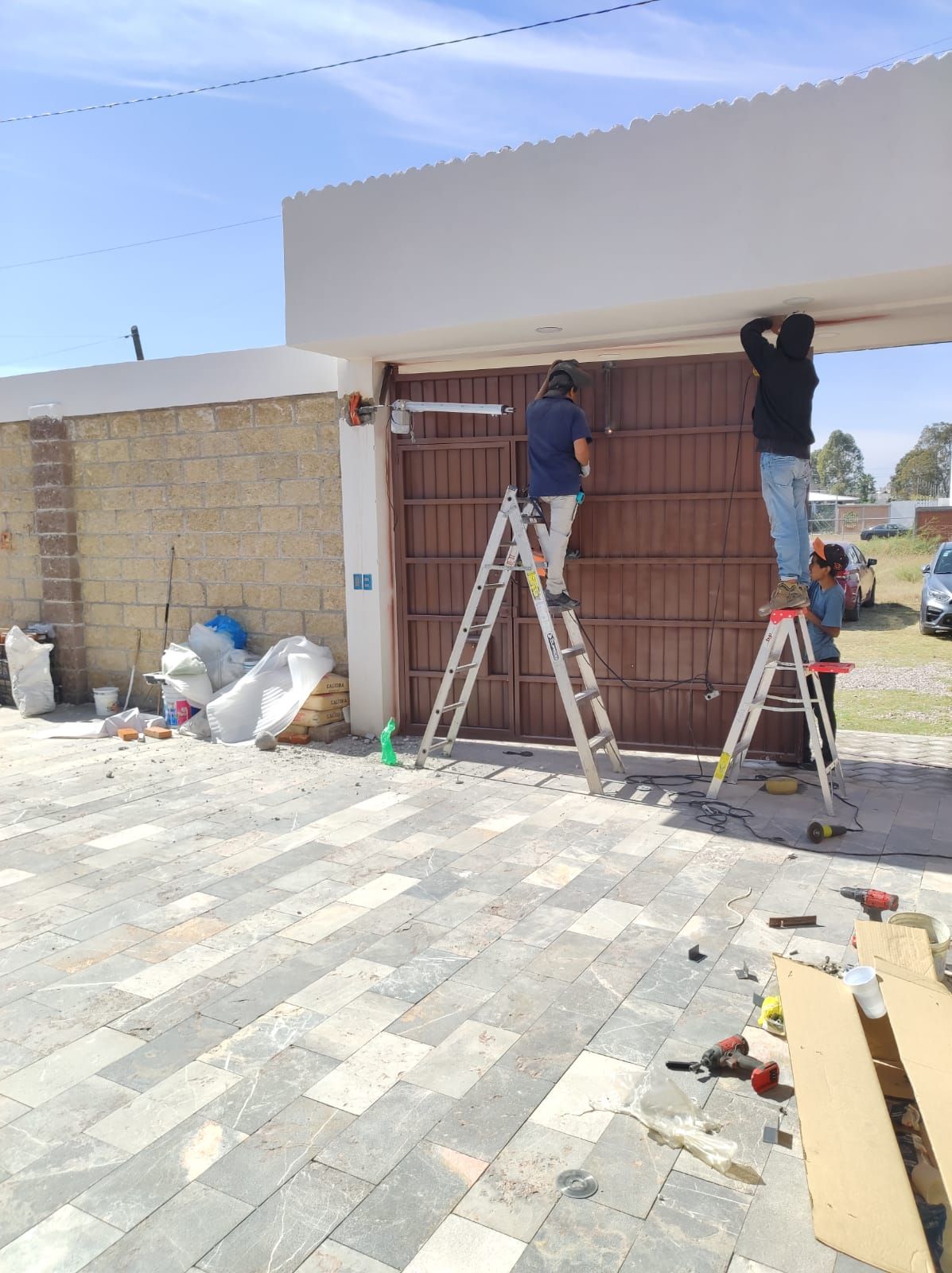 Dos hombres en escaleras instalando un motor en una puerta de metal marrón; pared de ladrillos, cielo azul.