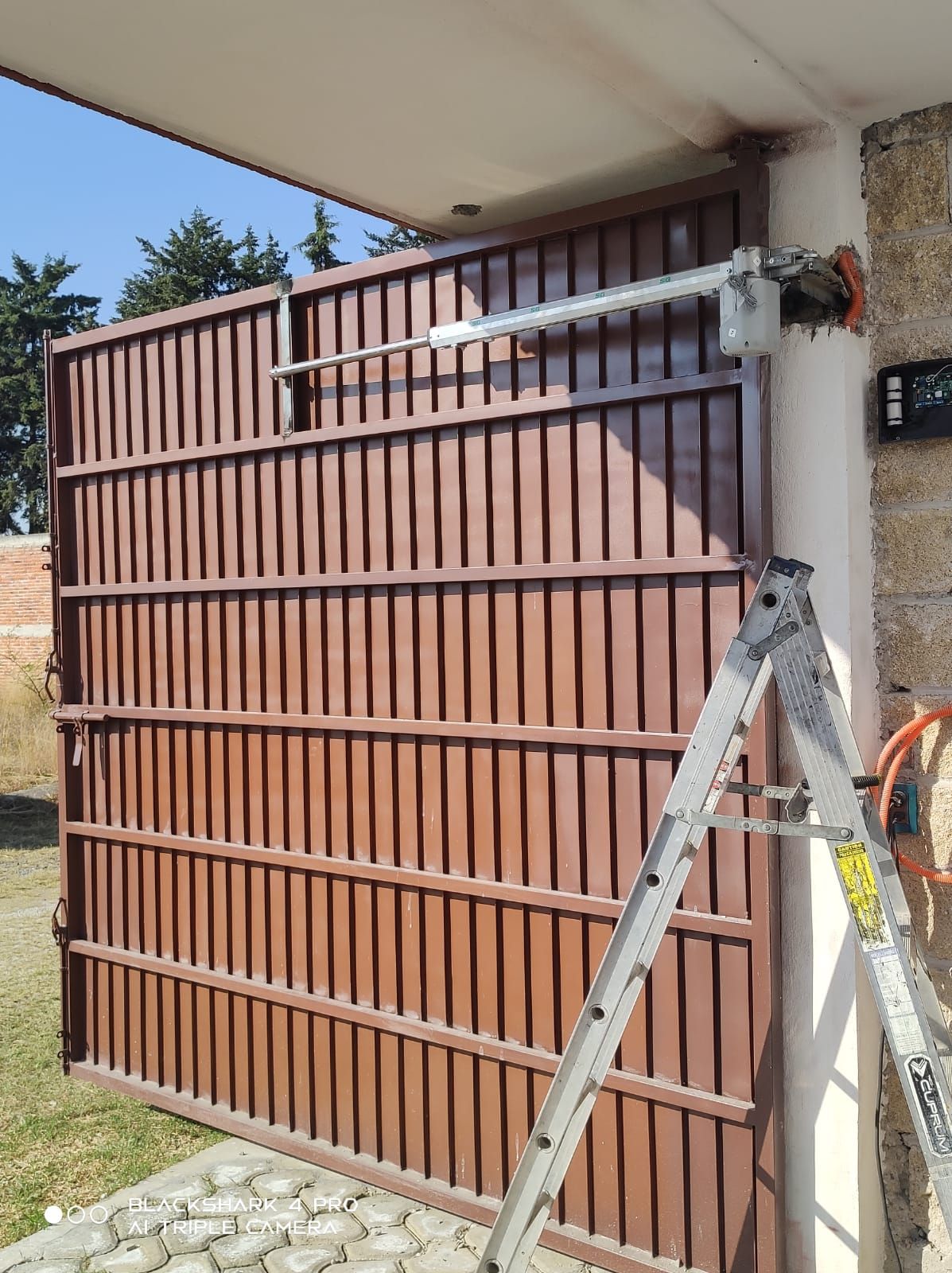Puerta metálica de color marrón con apertura eléctrica; hay una escalera apoyada contra la pared.