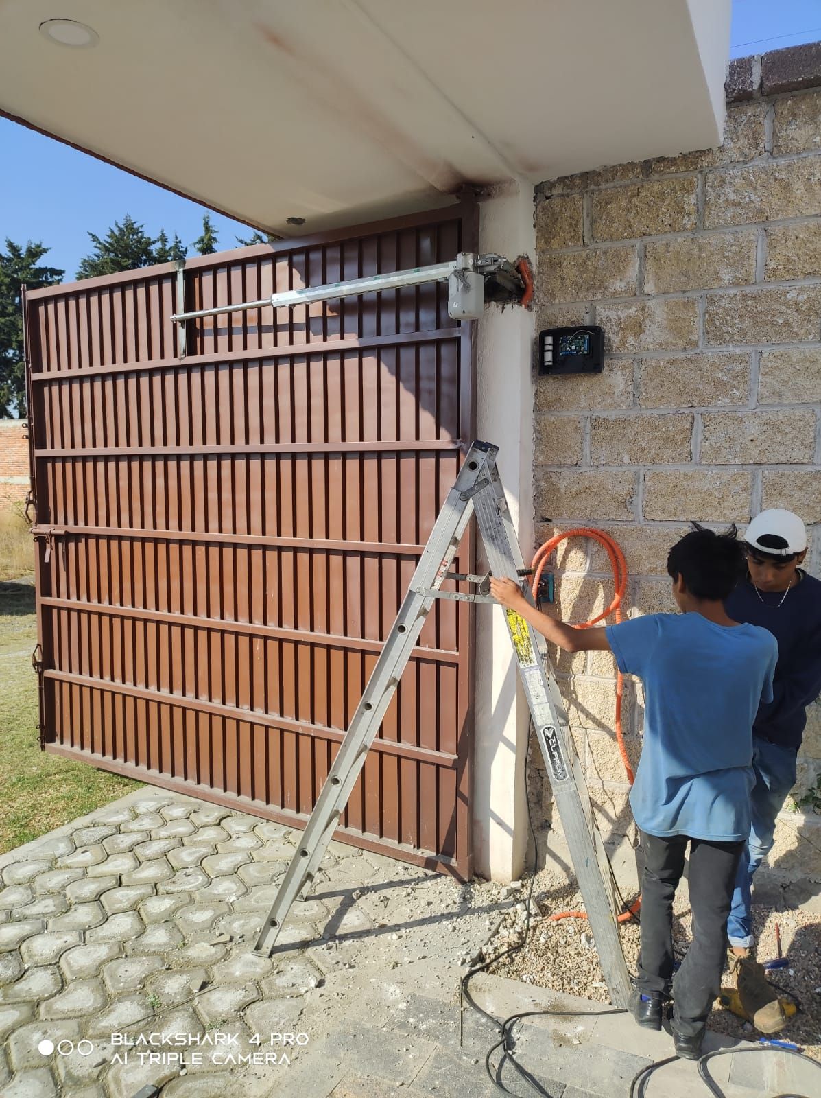 Dos personas instalan un abridor automático de portones en una puerta metálica marrón. Usan una escalera.