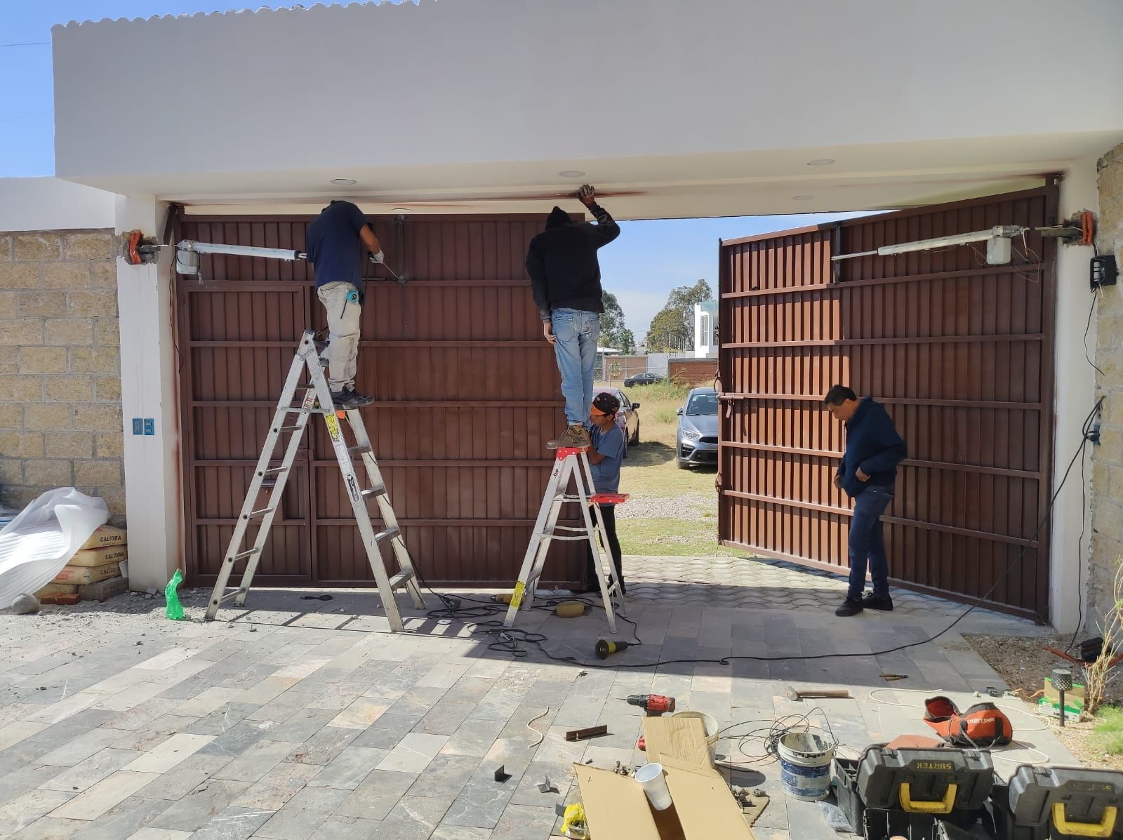 Trabajadores instalando un sistema de portón automático. Puerta metálica marrón, escaleras, herramientas y un exterior nublado.