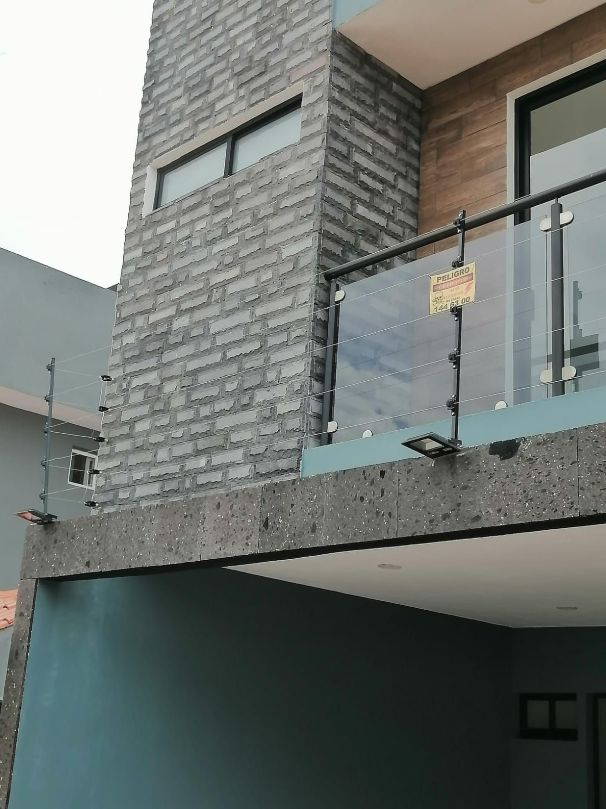 Exterior de casa de dos pisos, detalles de ladrillo gris, balcón de vidrio con cerca de seguridad y revestimiento azul grisáceo.