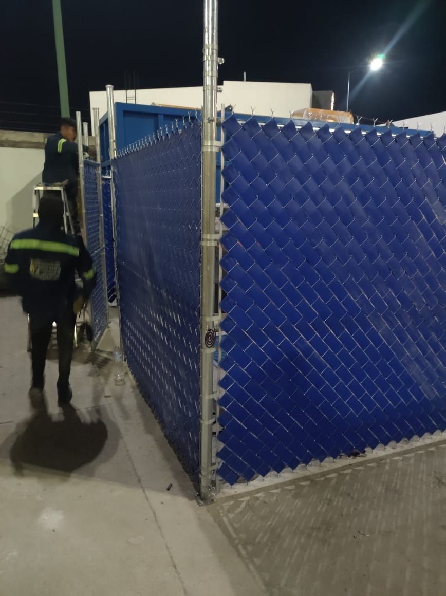 Dos trabajadores instalando una cerca azul de alambre al aire libre. Uno en una escalera, otro en el suelo. De noche.