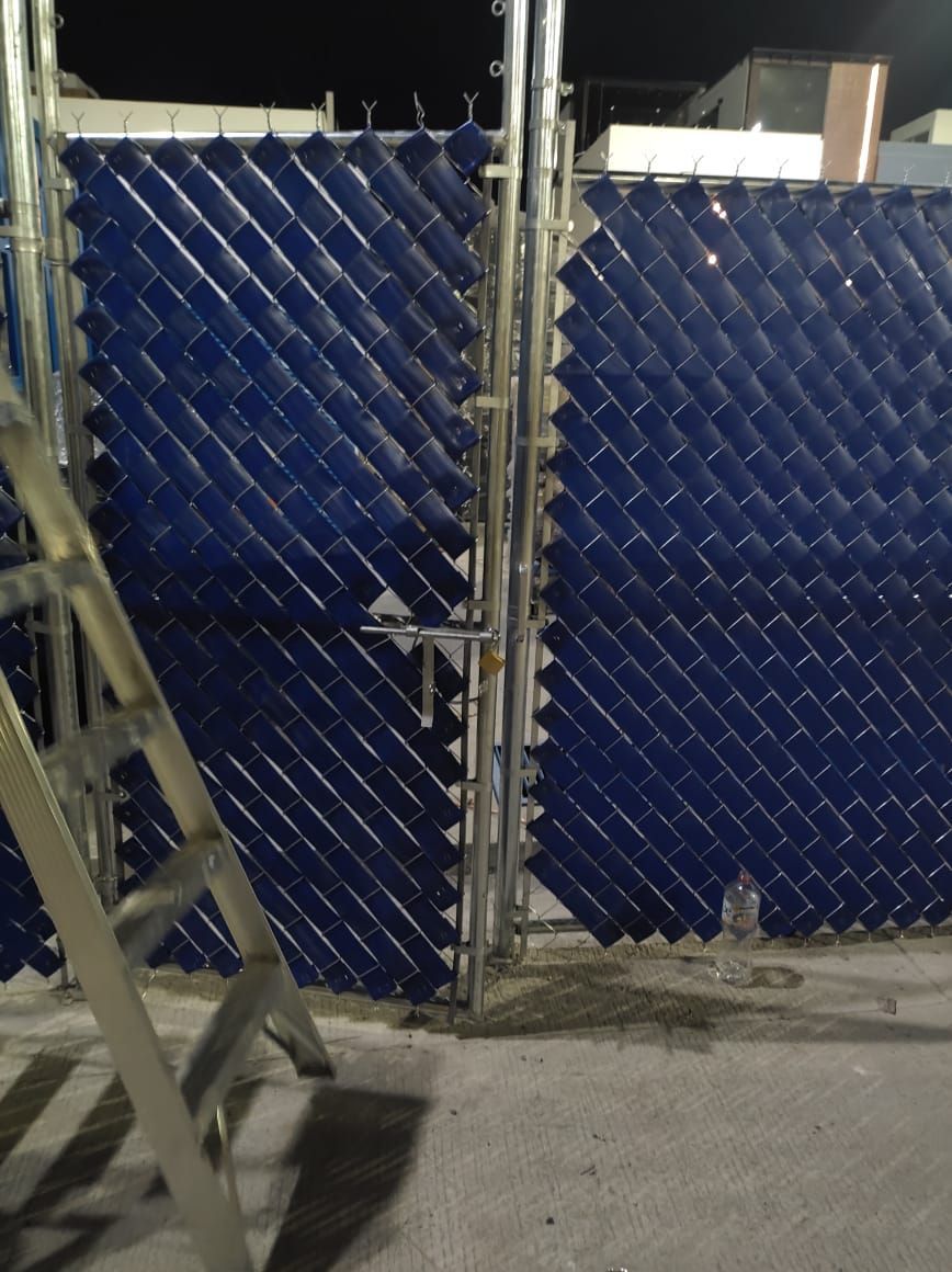 Puerta de malla azul parcialmente cubierta. Una escalera metálica se apoya en ella. Escenario nocturno.