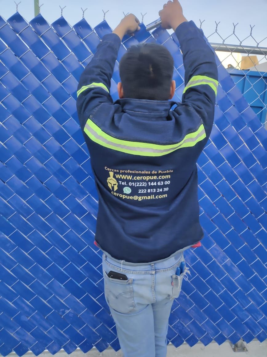 Una persona que viste una chaqueta azul está tejiendo listones de plástico azul a través de una cerca de alambre.