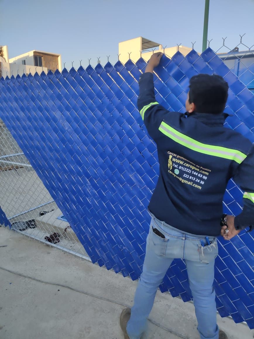 Hombre con chaqueta azul instalando láminas azules de privacidad en una cerca de alambre al aire libre.