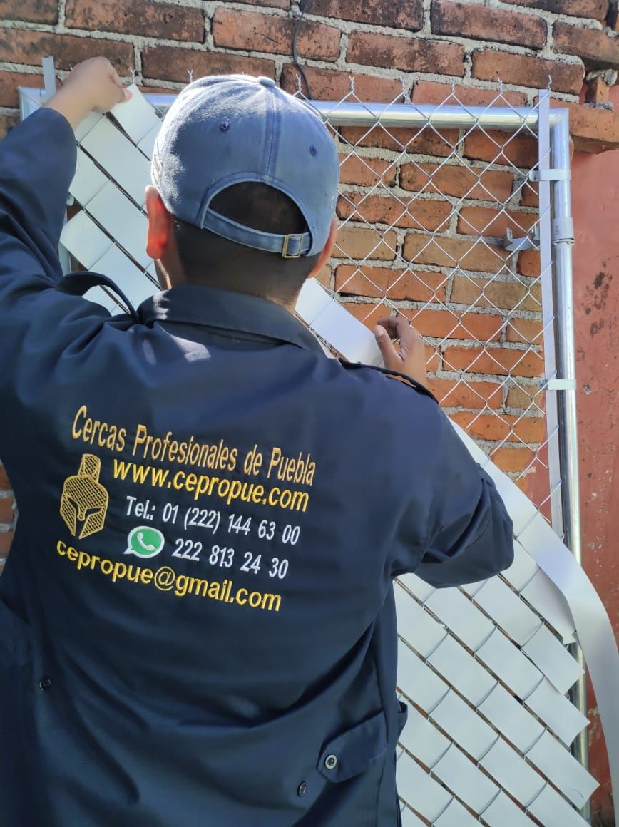 Persona instalando listones blancos de privacidad en una cerca de alambre cerca de un muro de ladrillo. Se ve un uniforme azul marino con el logotipo de la empresa.