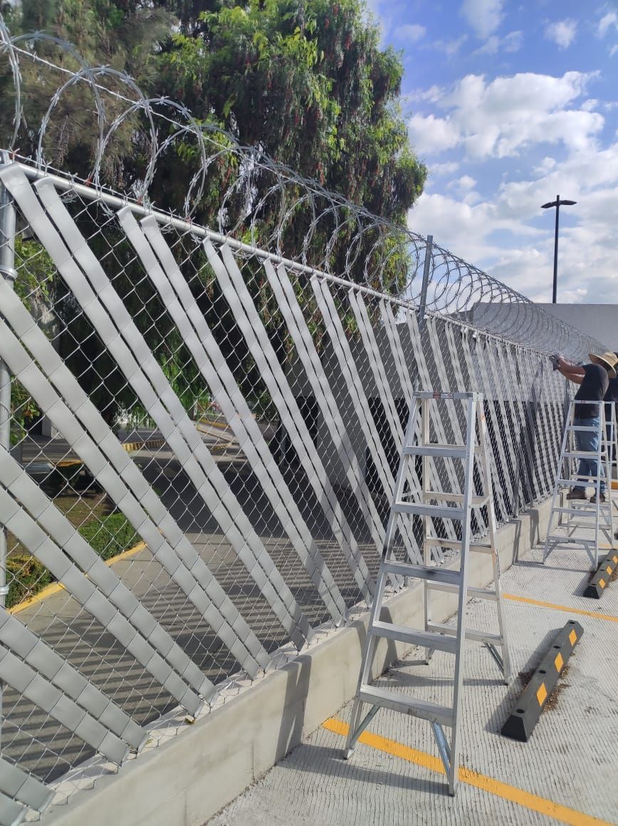 Valla de seguridad con alambre de púas, soportes metálicos y escalera en estacionamiento al aire libre.