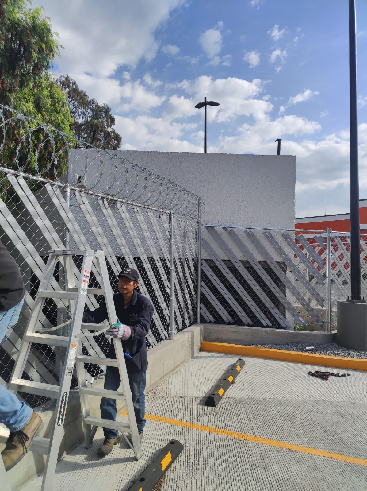 Persona en una escalera cerca de una valla de seguridad con alambre de púas, en un estacionamiento. Cielo nublado.