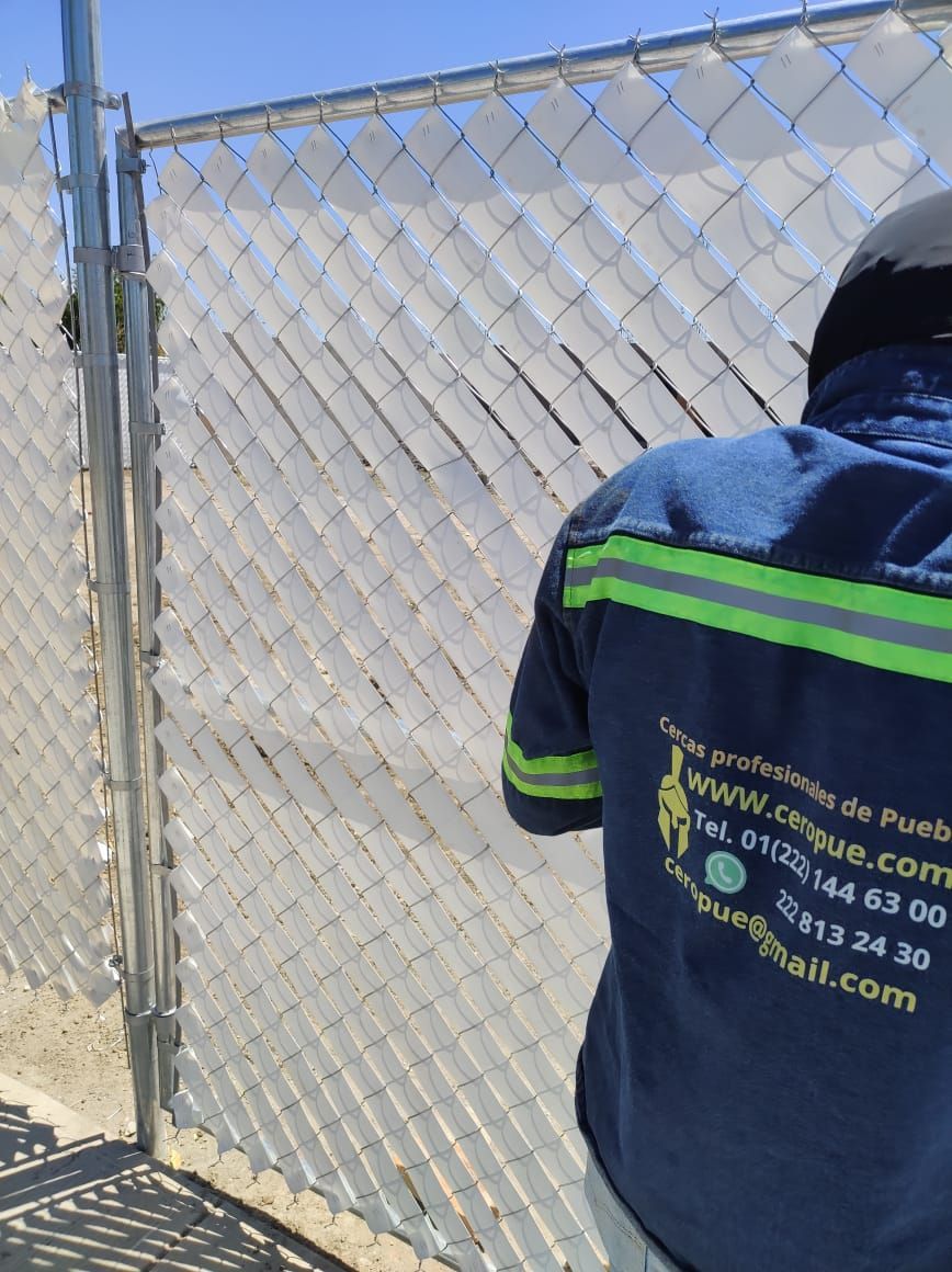 Un trabajador con uniforme azul instala láminas blancas para proteger la privacidad en una cerca de alambre.