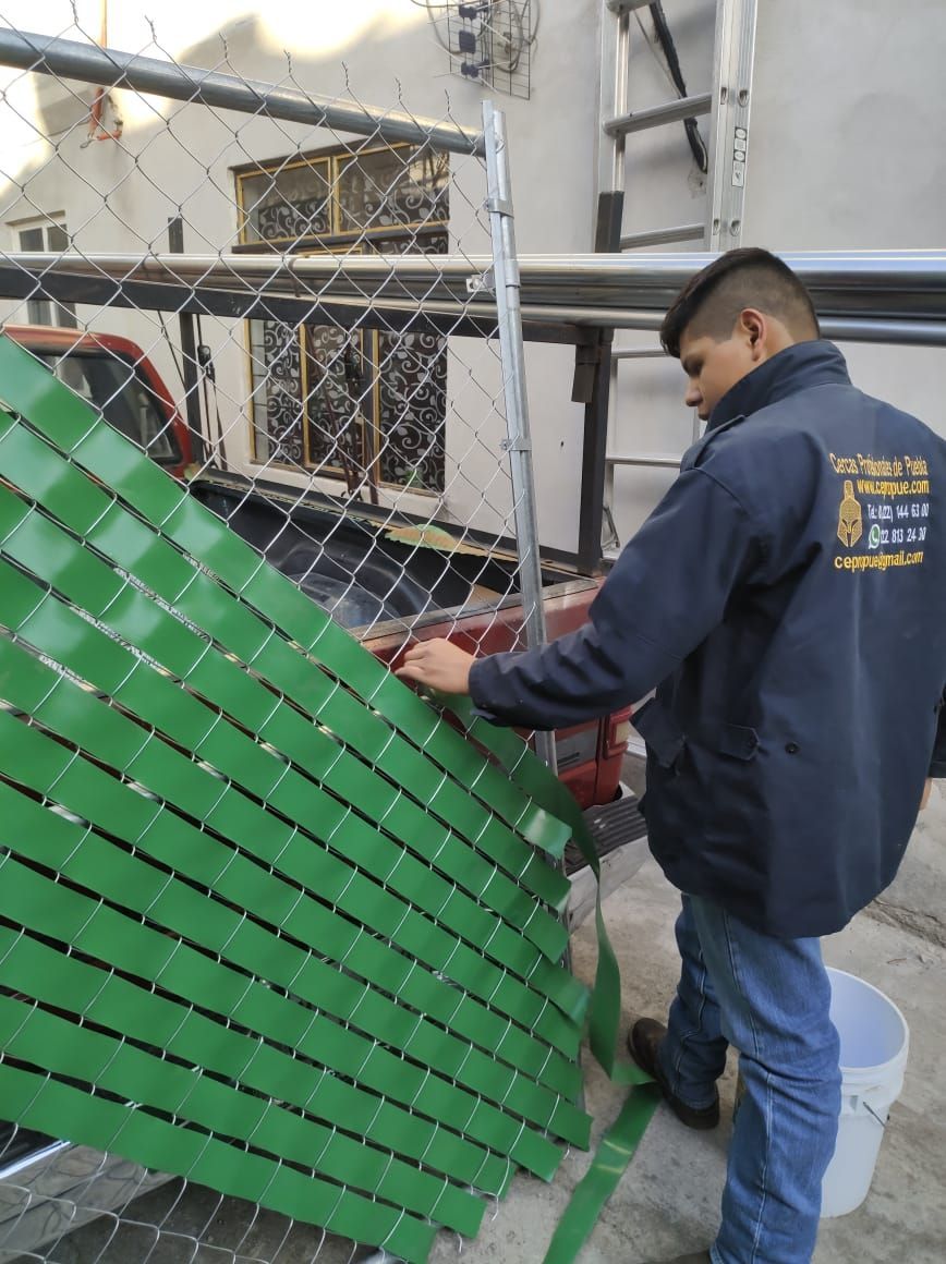 Hombre instalando láminas verdes para privacidad en una cerca de alambre en el exterior.