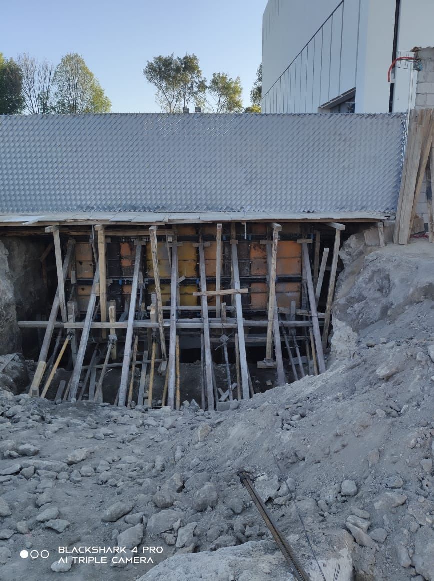 Sitio de construcción con estructura de madera que sostiene una estructura de hormigón; tierra, escombros y un edificio al fondo.