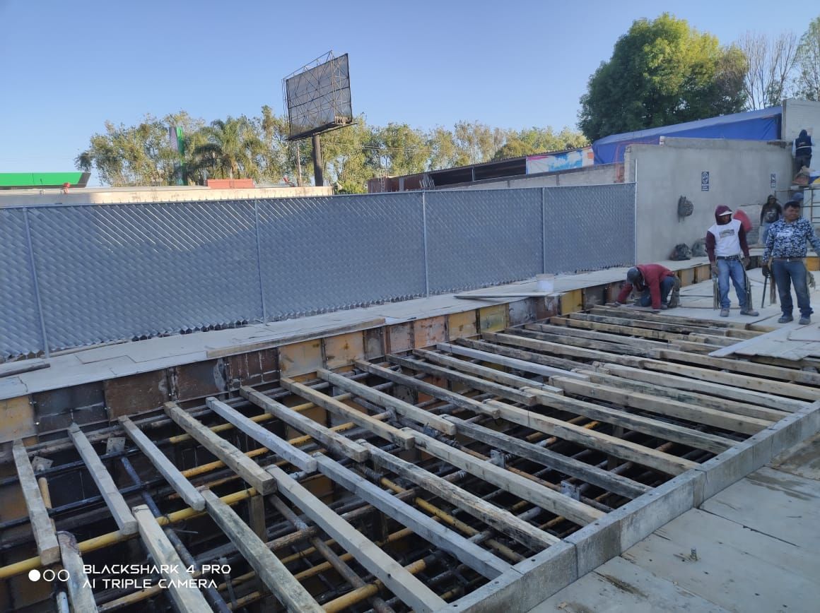 Sitio de construcción con trabajadores; andamios de madera, valla metálica, hormigón gris, árboles al fondo.