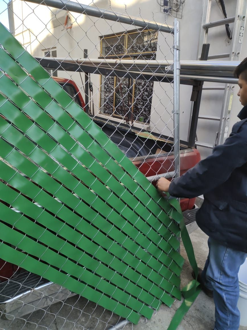 Hombre instalando láminas verdes para privacidad en una cerca de alambre en el exterior.