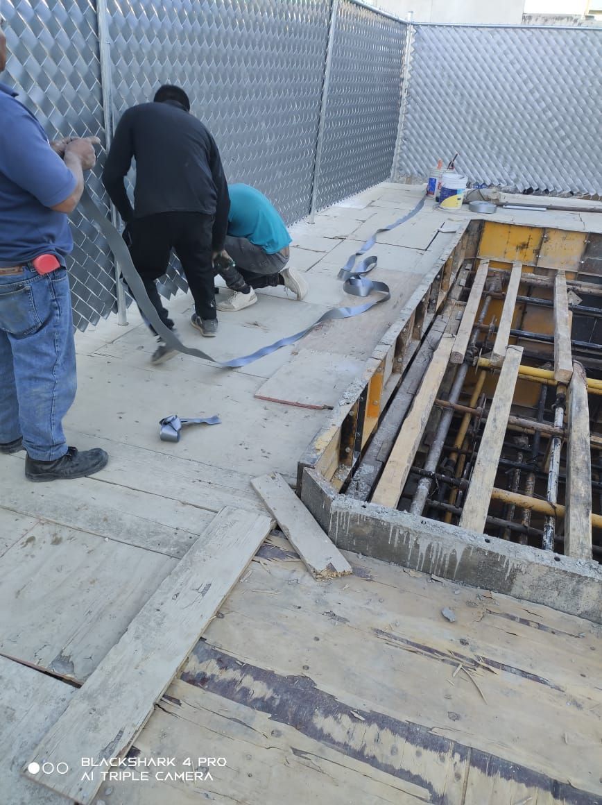 Obreros de la construcción en una plataforma de madera, formando una estructura de hormigón. Valla metálica al fondo.