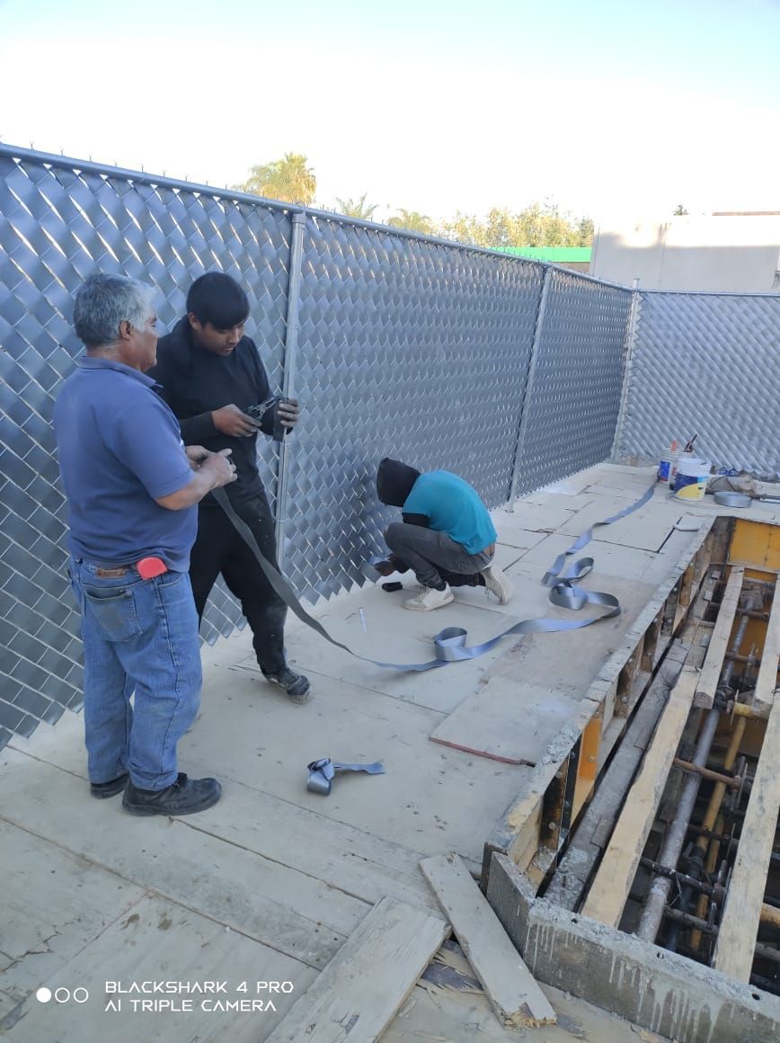 Tres obreros de la construcción cerca de una valla metálica y una estructura de madera. Uno se arrodilla, dos se ponen de pie e inspeccionan.