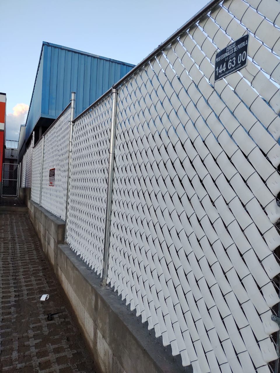 Valla de alambre cubierta de blanco a lo largo de una pasarela de ladrillos, con un edificio azul visible al fondo.