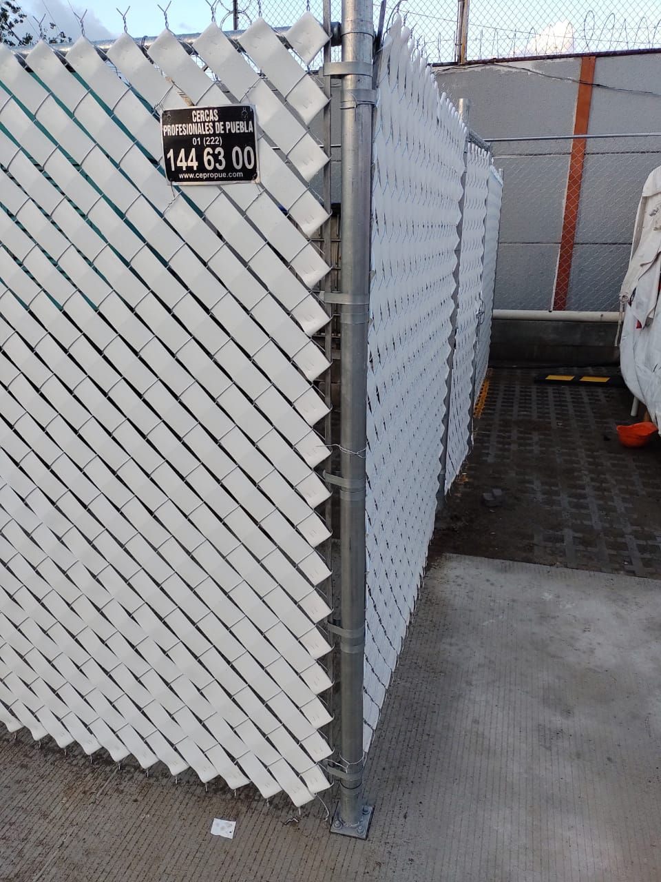 Valla de privacidad blanca con listones diagonales; se ve un poste de metal.