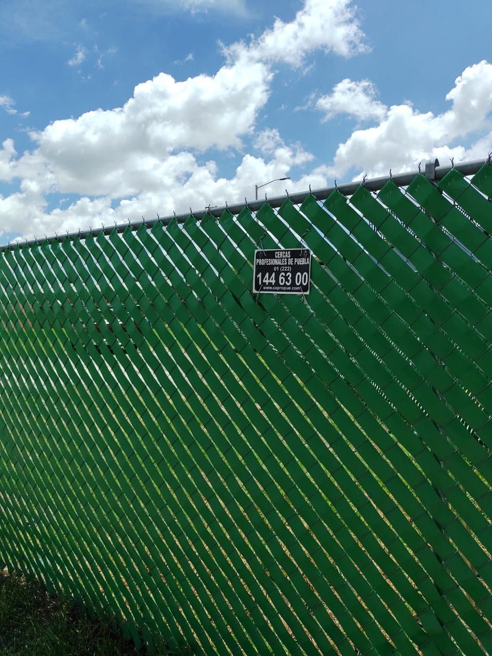 Valla de alambre verde con listones verdes. Un pequeño letrero está fijado a la valla. Cielo azul y nubes al fondo.