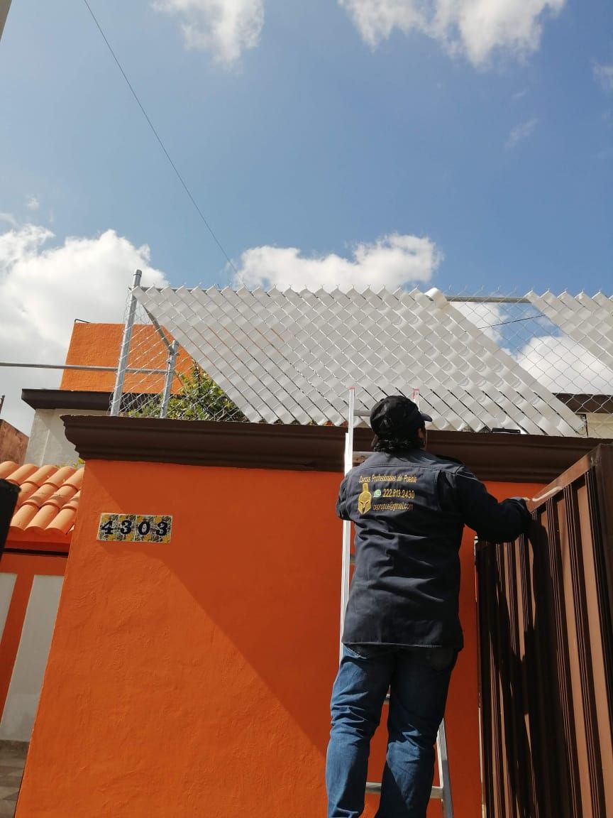 Un trabajador en una escalera cerca de una pared y una puerta de color naranja, instalando una cerca de alambre en un techo.