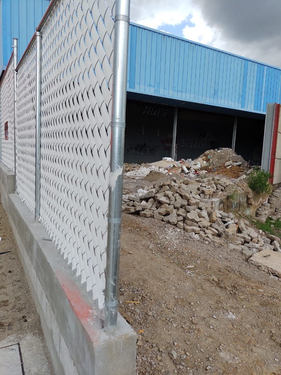 Valla de alambre con insertos blancos delante de un edificio con techo azul, tierra y escombros en el frente.