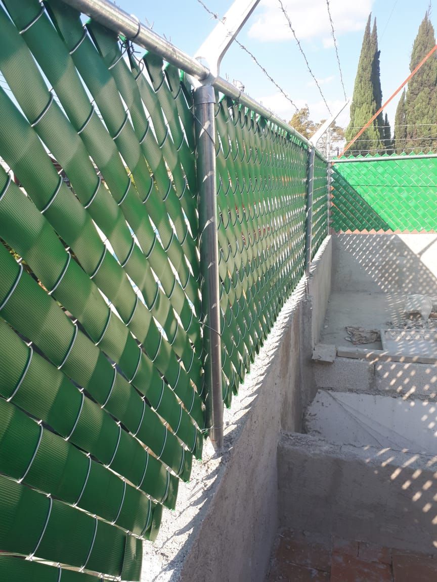 Valla de alambre de cadena verde con listones de privacidad y alambre de púas sobre una pared de hormigón, día soleado.