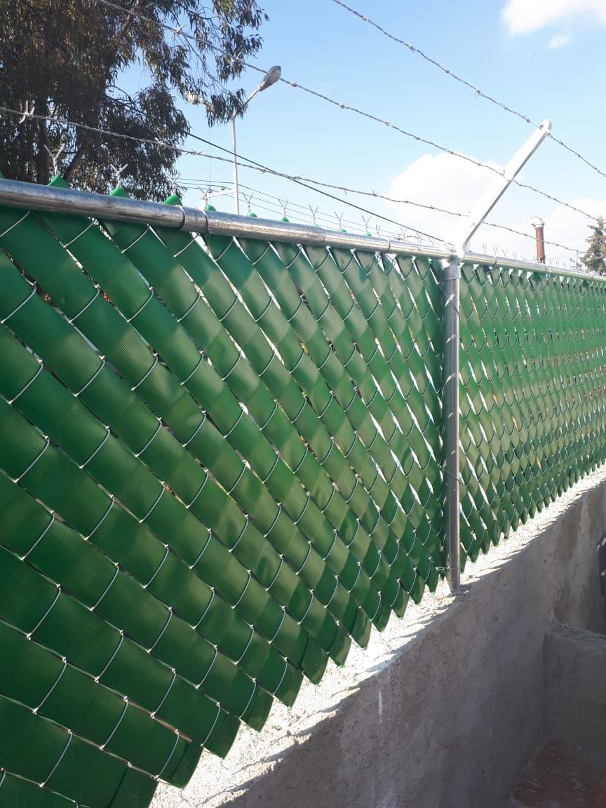 Valla de alambre verde con listones verdes y alambre de púas en la parte superior, contra un cielo azul.