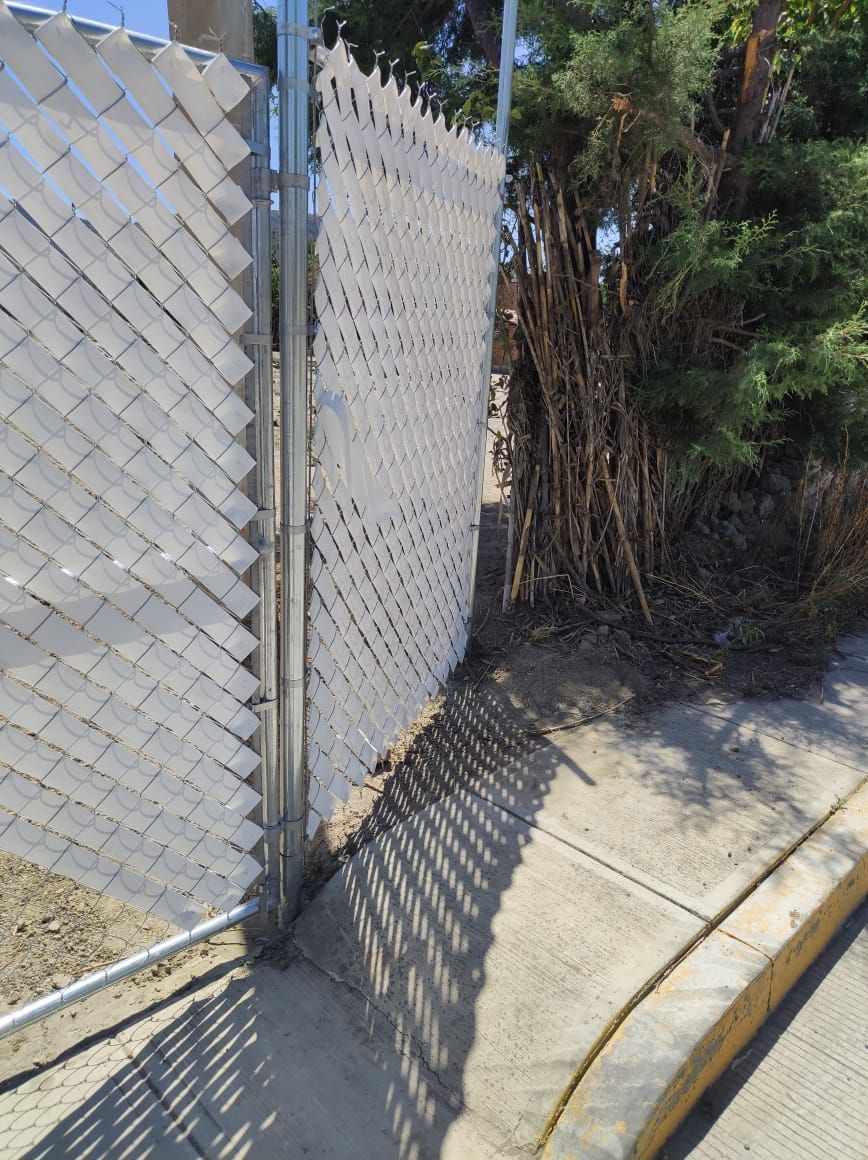 Valla de alambre con listones blancos para privacidad. Puerta abierta y acera adyacente.