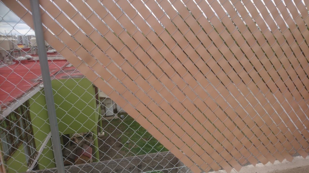 Listones de color canela tejidos en una cerca de alambre, ocultando parcialmente un edificio y una pared verde.