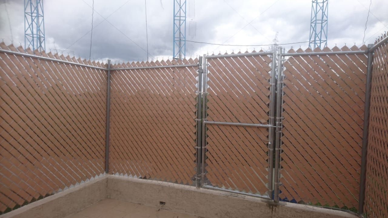 Valla de celosía marrón con puerta, que rodea un área de hormigón, con líneas eléctricas al fondo.